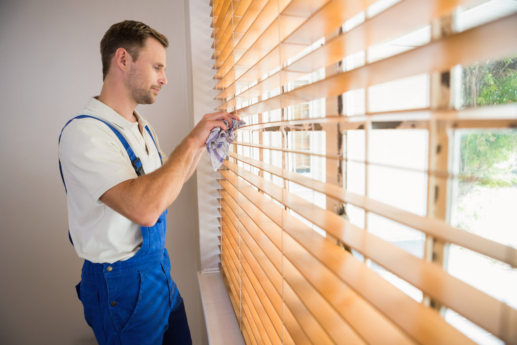 Clear Choice: A man in a blue uniform and overalls dusting wooden blinds beside a bright window in Boise Valley.