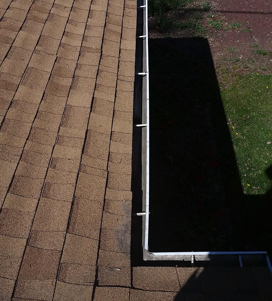 Clear Choice: Aerial view of a brown shingled roof with a white gutter cleaning system beside a garden with green grass and red flowers in the Boise Valley.