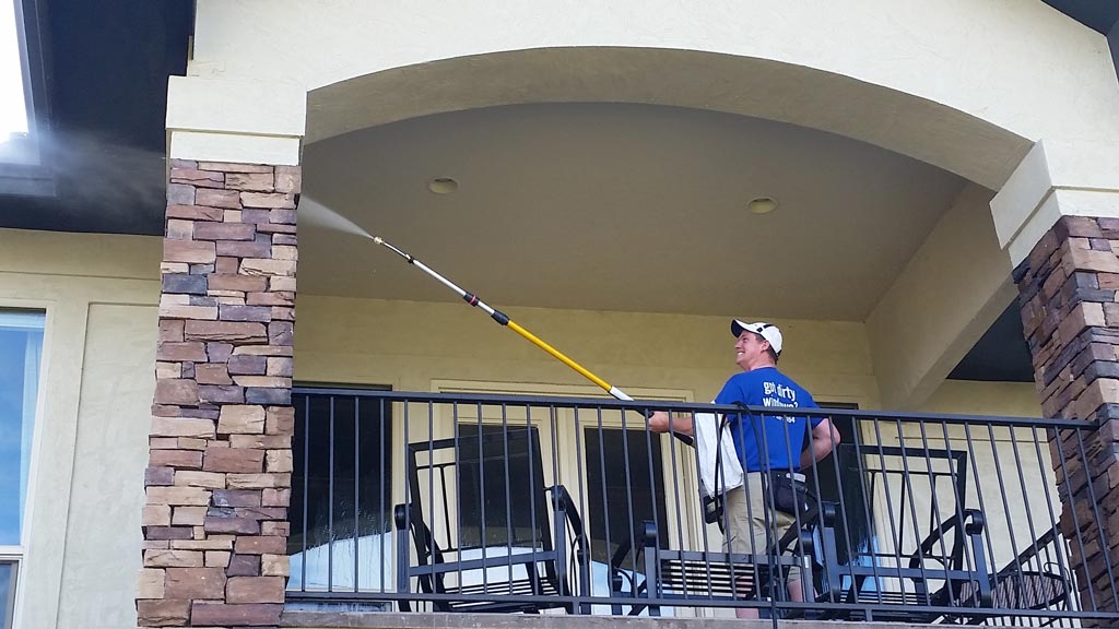 Clear Choice: A man in a blue shirt and cap using a long extendable brush to power wash the ceiling of a balcony on a stone and stucco building.