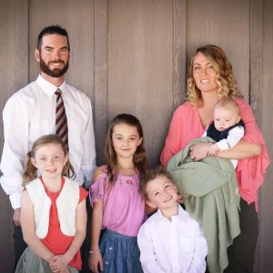 Clear Choice: A family of six poses together, including a man, woman, three young girls, and a baby held by the woman, standing against a wooden wall.