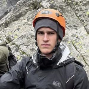 Clear Choice: Person wearing an orange climbing helmet, gray beanie, and black jacket stands beside a rocky surface.