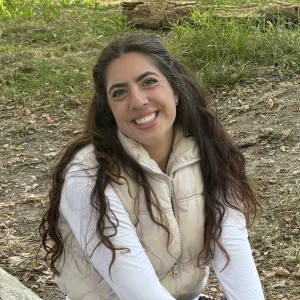 Clear Choice: Person with long hair smiling outdoors, wearing a light vest and white shirt, sitting on grass with fallen leaves around.