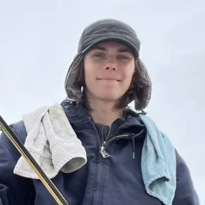 Clear Choice: A member of Our Team stands outdoors against a grey sky, wearing a dark jacket and hat, with two towels draped over their shoulders.