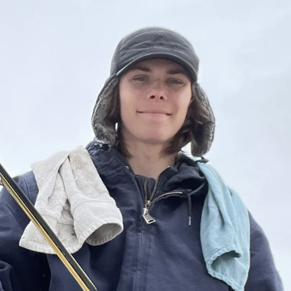 Clear Choice: A member of Our Team stands outdoors against a grey sky, wearing a dark jacket and hat, with two towels draped over their shoulders.