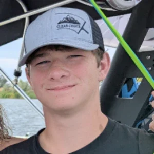 Clear Choice: A teenage boy wearing a gray and black hat with a mountain logo stands outdoors, with water and trees visible in the background.