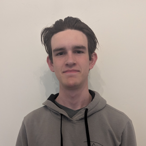 Clear Choice: A young man with short brown hair stands in front of a plain off-white wall, wearing a gray hoodie and a neutral expression, as if waiting to join the organization.