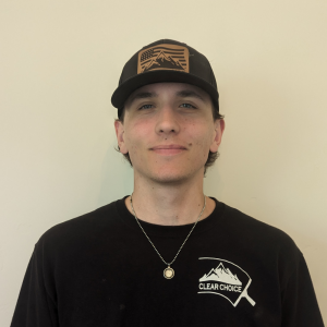 Clear Choice: A member of our team is wearing a black "Clear Choice" t-shirt and black cap, standing against a plain background. The cap features a mountain design, and they are also sporting a necklace.