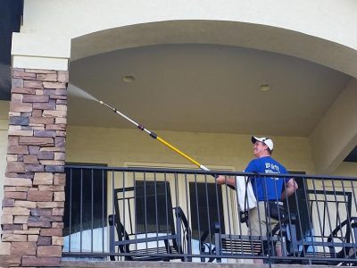 Clear Choice: A man in a blue shirt and cap using a long extendable brush to power wash the ceiling of a balcony on a stone and stucco building.
