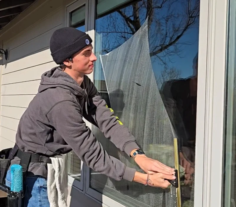 Clear Choice: Person cleaning a house window with a squeegee, standing on a step ladder, wearing a beanie and hoodie. Cleaning supplies are attached to their belt.